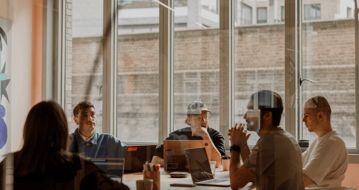 Team meeting in modern office with large windows and laptops on a table, focused on discussion and collaboration.