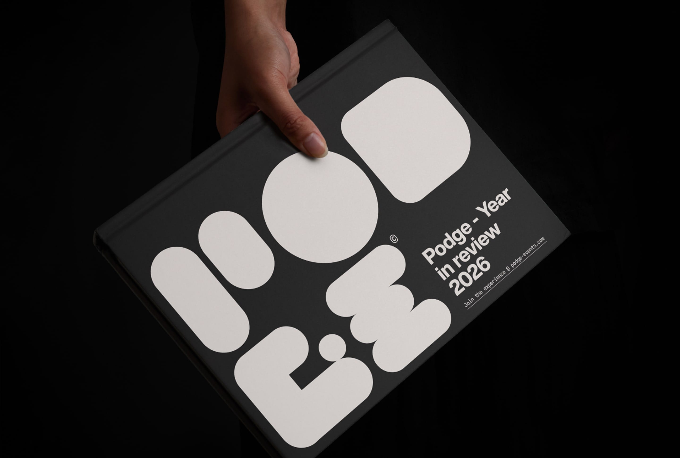 Person holding Podge - Year in Review 2026 book with bold black and white design, against a dark background.