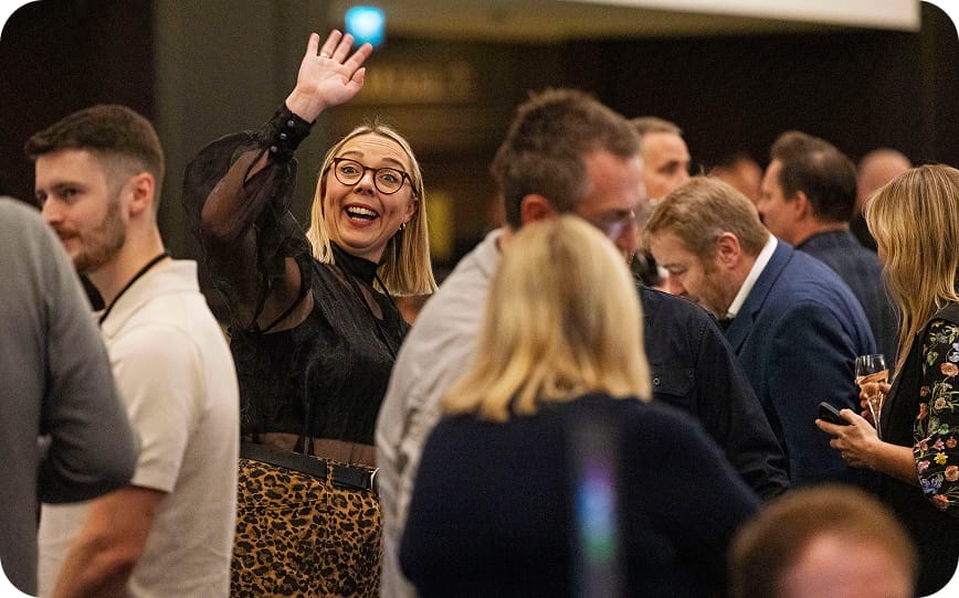 A cheerful woman waves amidst a lively networking event, engaging with the crowd.